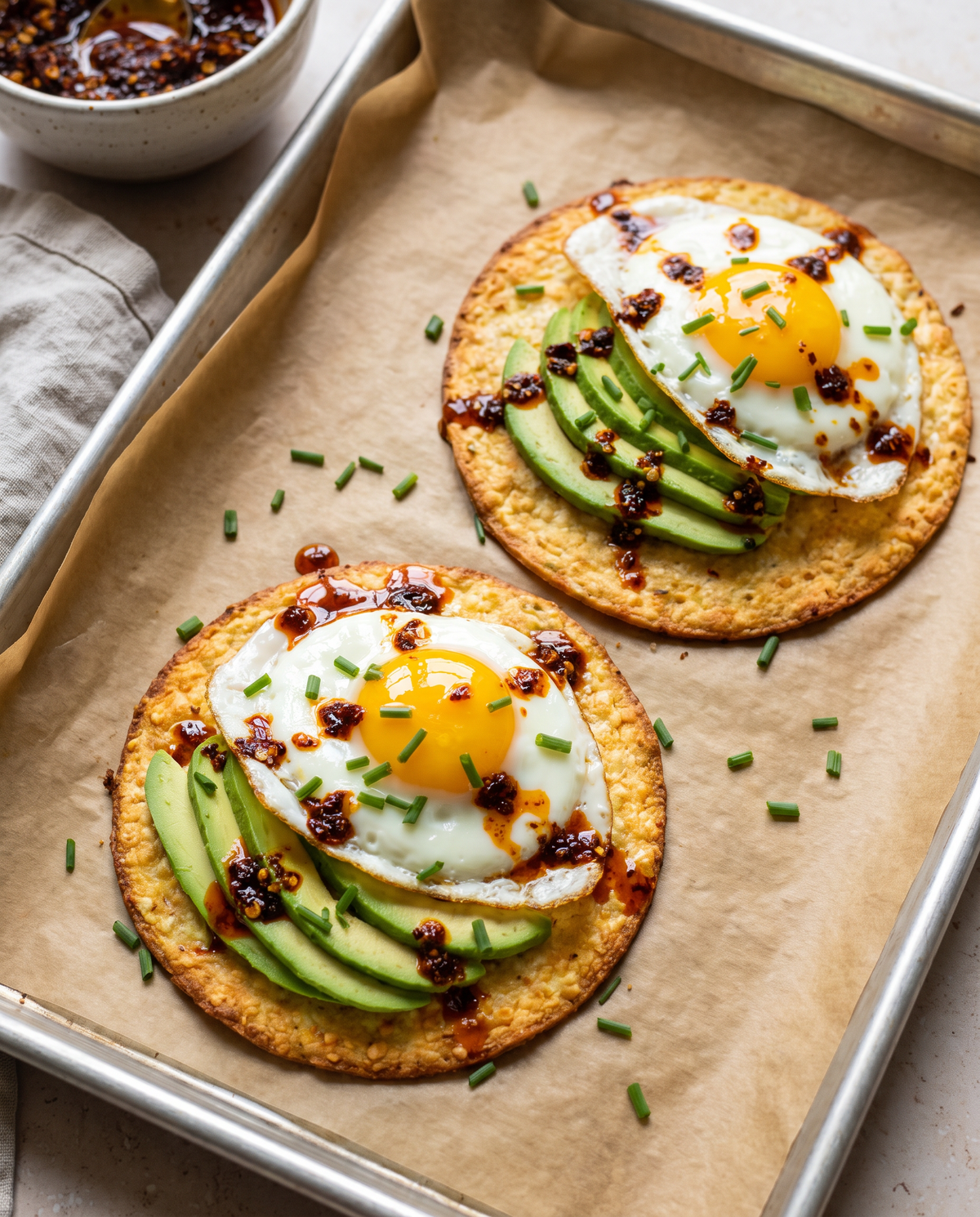 Two baked cottage cheese taco shells topped with avocado, fried eggs, chili oil and chives on parchment.